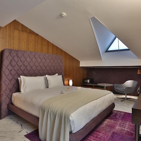 Dosso Dossi Hotels & Spa Downtown Estambul Exterior foto The photo shows a stylish hotel room with a modern design. It features a large bed with a tufted purple headboard, complemented by white bedding and a light gray throw. The walls are made of wooden panels, adding warmth to the space. To the side, the