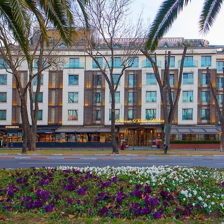 Dosso Dossi Hotels & Spa Downtown Estambul Exterior foto The photo shows a building with a modern design, featuring a facade with large windows and a mix of materials. In the foreground, there are colorful flower beds with purple and white flowers. The scene is framed by tall trees, and the overall setting
