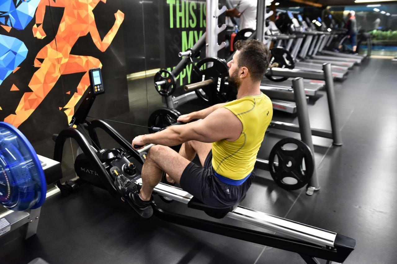 Dosso Dossi Hotels & Spa Downtown Estambul Exterior foto The photo shows a man exercising on a rowing machine in a gym. He is wearing a yellow tank top and black shorts. The background features an exercise-themed mural with colorful graphics of a person in motion. Various gym equipment, including treadmill