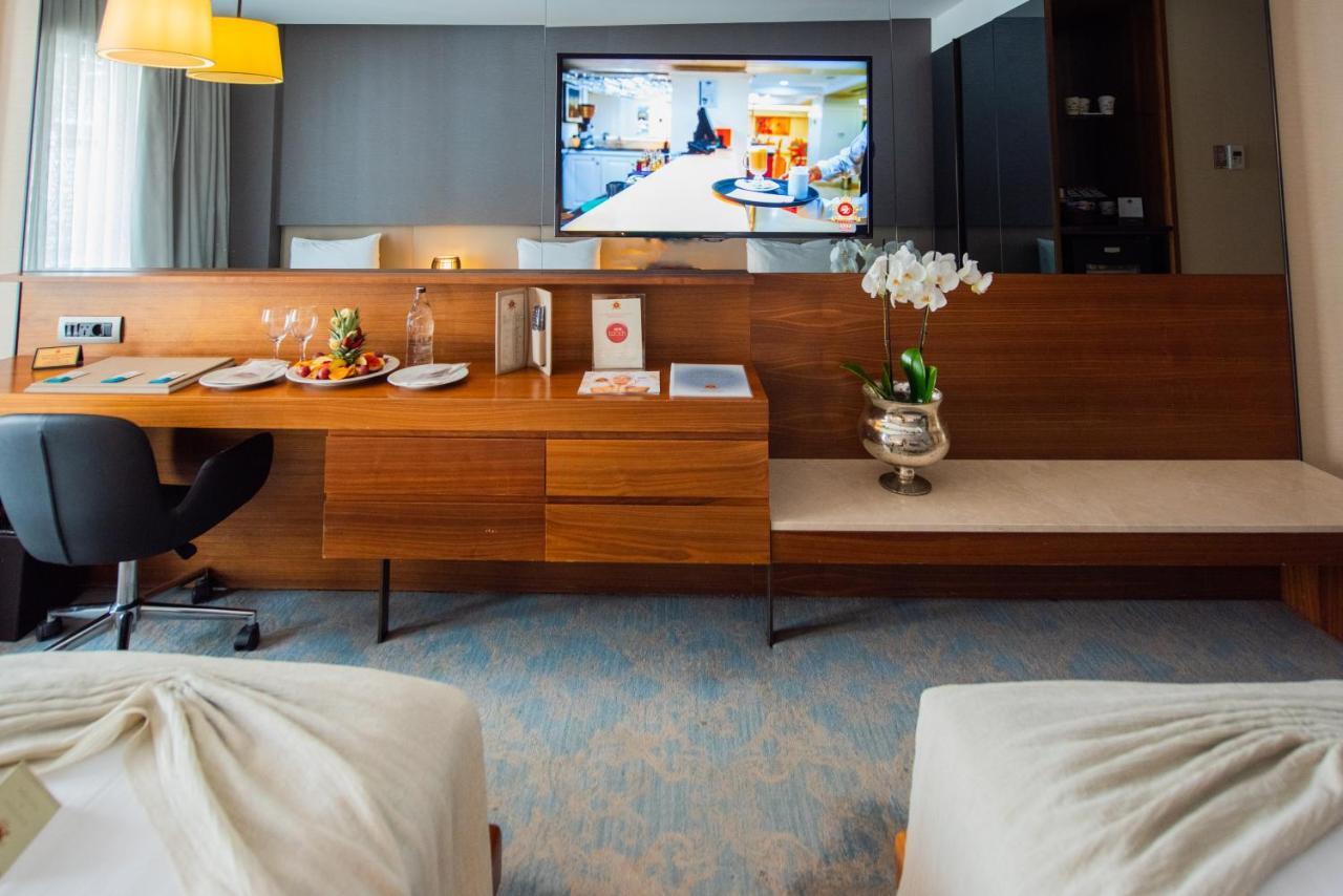 Dosso Dossi Hotels & Spa Downtown Estambul Exterior foto The photo shows a modern hotel room featuring two beds with light-colored bedding. In the background, there is a flat-screen television mounted on the wall displaying what appears to be a hotel service or entertainment program. A wooden console table