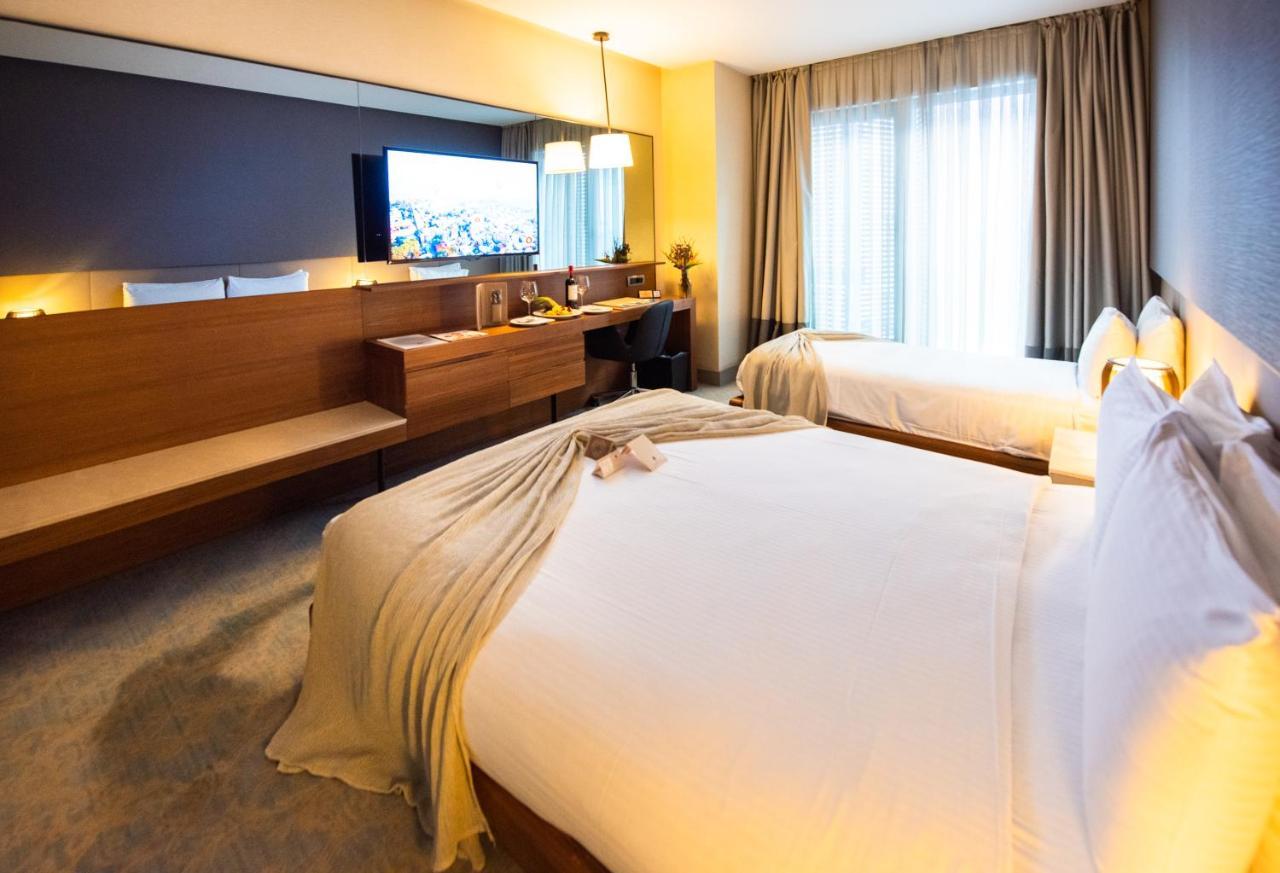 Dosso Dossi Hotels & Spa Downtown Estambul Exterior foto The photo shows a modern hotel room featuring two beds with white linens and a decorative throw blanket. There's a wooden desk and a chair alongside a large flat-screen TV mounted on the wall. The room has large windows with curtains, allowing natura