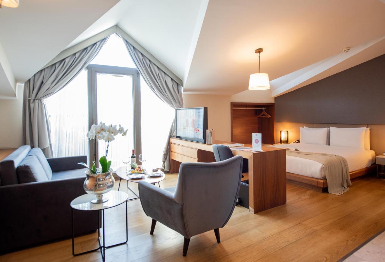 Dosso Dossi Hotels & Spa Downtown Estambul Exterior foto The photo shows a stylish hotel room with a modern design. It features a comfortable bed in one area, complemented by a cozy seating area with a sofa and a chair. There is a small round table with a vase of flowers and some snacks. A flat-screen TV i