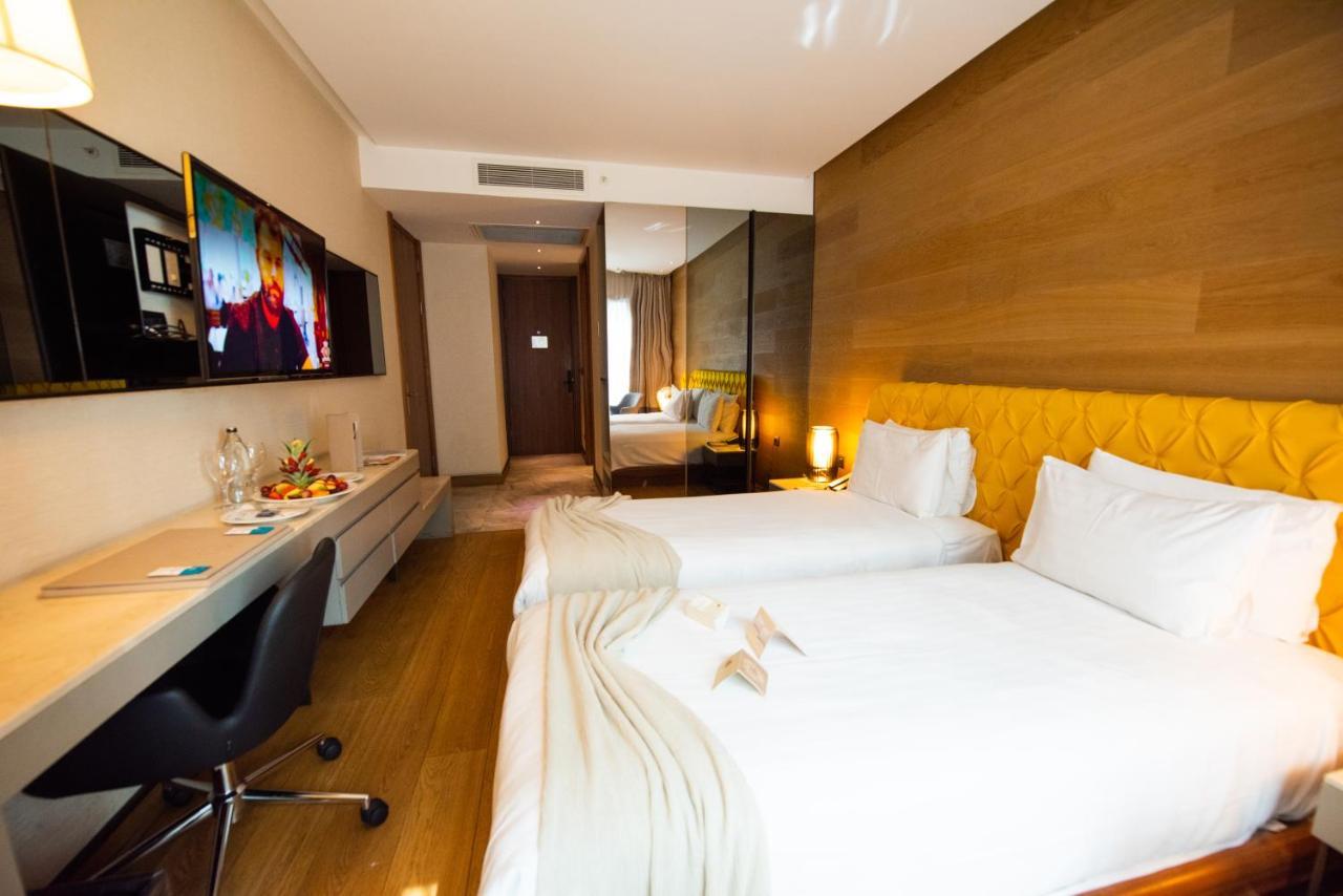 Dosso Dossi Hotels & Spa Downtown Estambul Exterior foto The photo shows a modern hotel room featuring two single beds with white linens. There is a stylish yellow accent wall behind the beds. A flat-screen television is mounted on the wall opposite the beds, and a sleek desk and chair are positioned next 