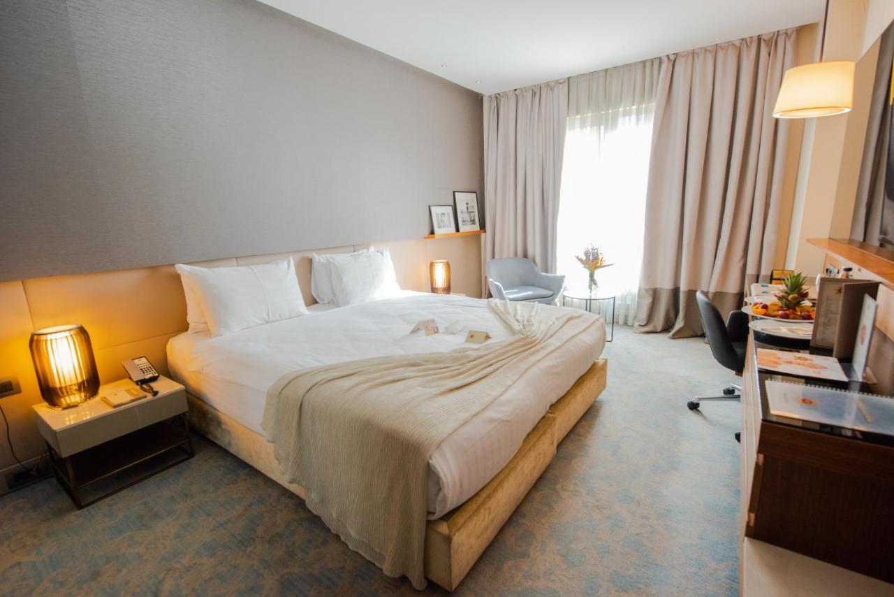 Dosso Dossi Hotels & Spa Downtown Estambul Exterior foto The photo showcases a modern hotel room featuring a king-sized bed with white bedding and a light-colored throw. There are elegant bedside lamps on either side of the bed and a small nightstand. A large window dressed with soft, light-colored curtain