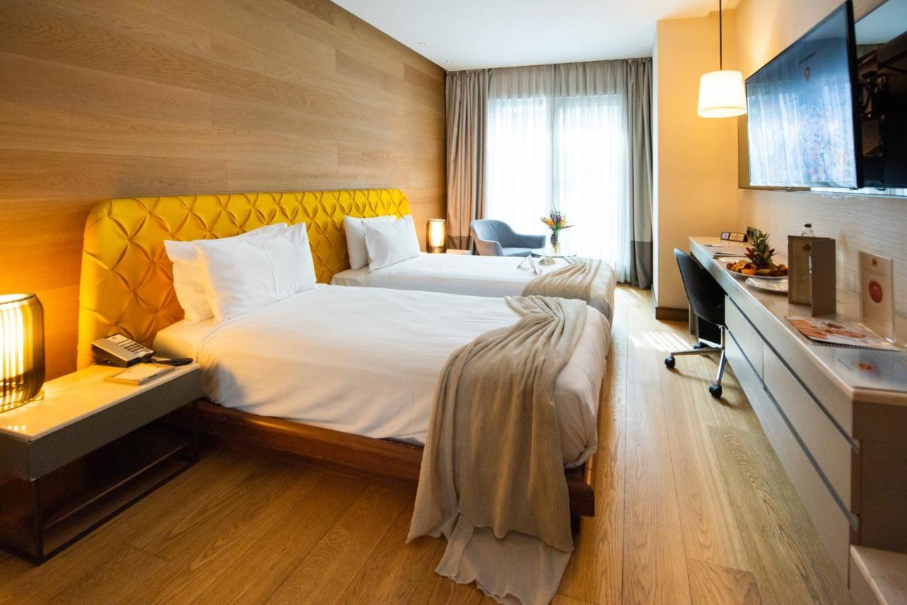 Dosso Dossi Hotels & Spa Downtown Estambul Exterior foto The photo shows a stylish hotel room featuring two beds with white linens and a unique yellow headboard. The room has wooden walls and a polished wooden floor. There are large windows with sheer curtains allowing natural light to flood in. A small se