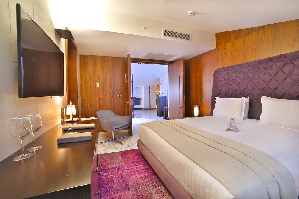 Dosso Dossi Hotels & Spa Downtown Estambul Exterior foto The photo depicts a modern hotel room. It features a large bed with white linens and a decorative headboard in a rich purple hue. The room has wooden paneling on the walls, adding warmth to the space. There's a flat-screen TV mounted on the wall oppo