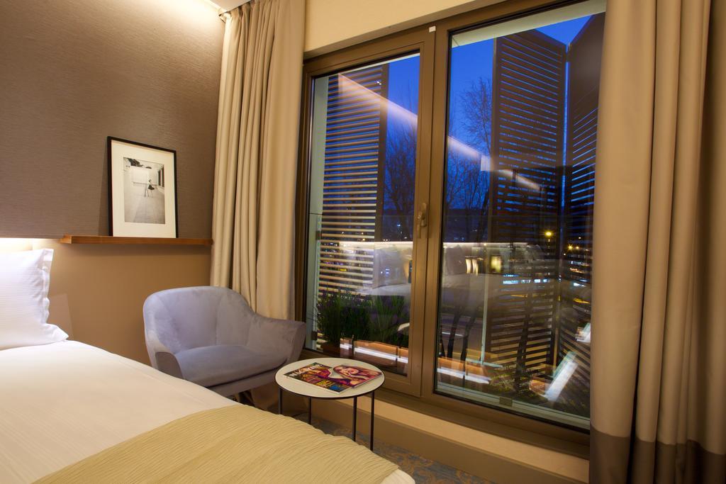 Dosso Dossi Hotels & Spa Downtown Estambul Exterior foto The photo depicts a modern hotel room. On the left side, there is a neatly made bed with a light-colored blanket. Next to the bed, there's a small round table with some magazines on top. A cozy chair in a light shade is positioned nearby. The main fe